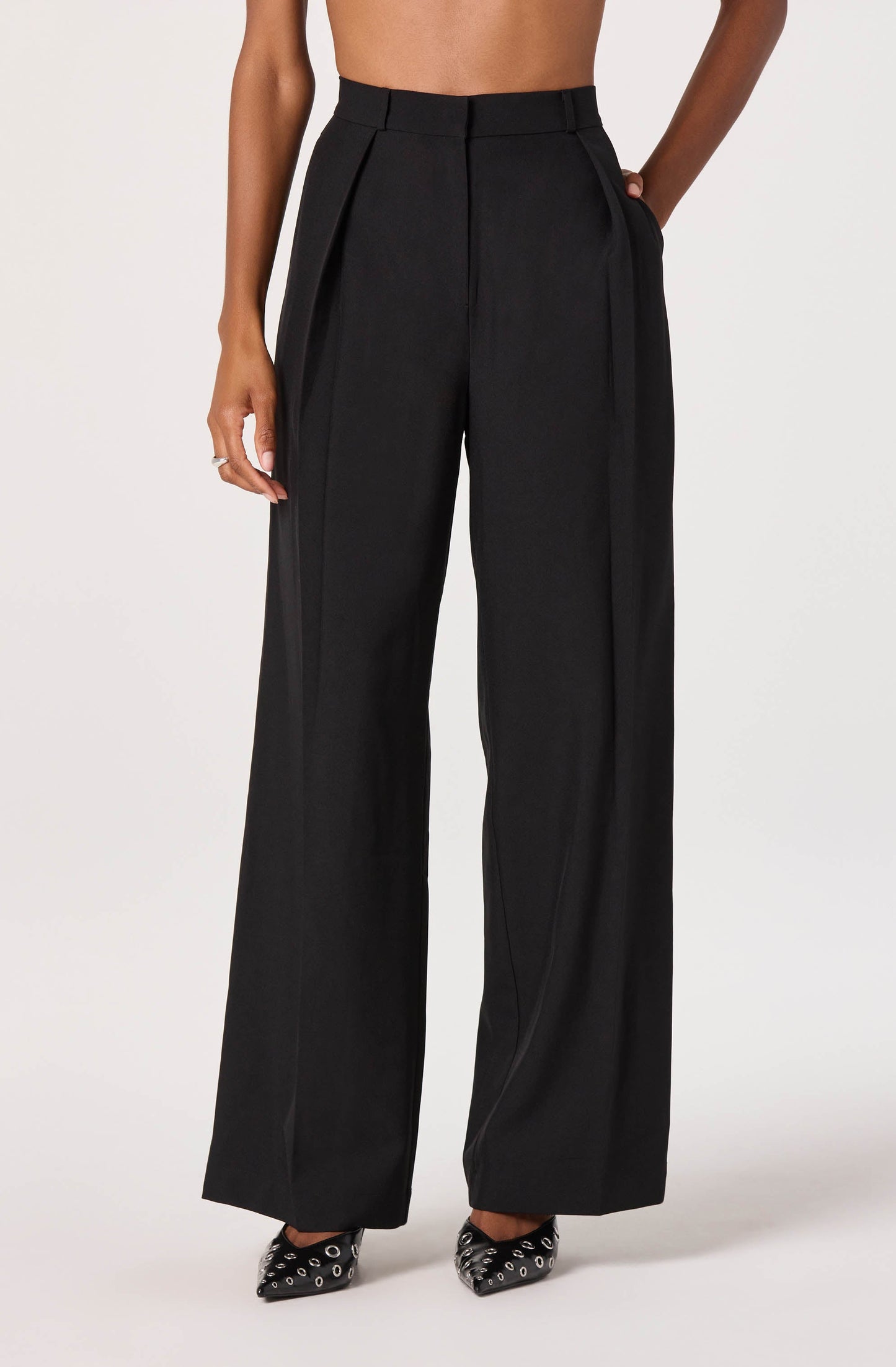 A person is wearing high-waisted black wide-leg trousers with pleats at the front, made of a smooth, slightly shiny fabric. The trousers have belt loops at the waistband and side pockets. The person is also wearing black pointed-toe high heels with decorative metallic eyelets.