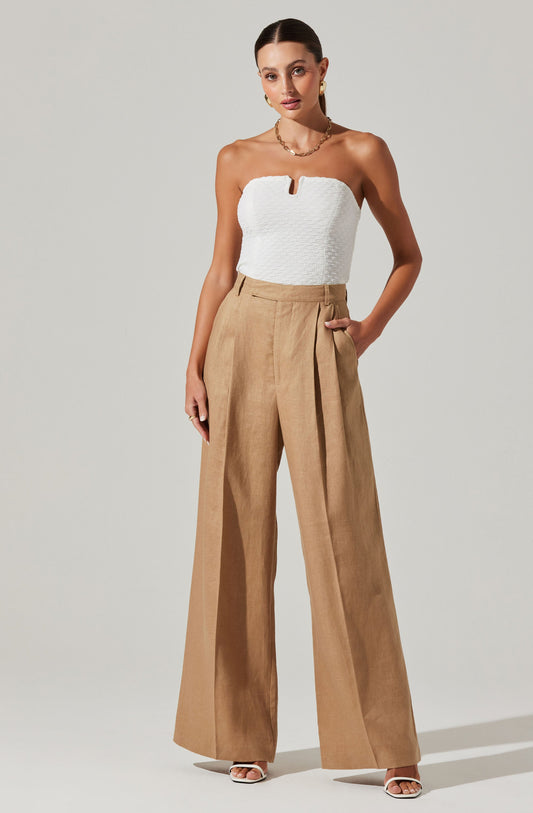 A woman is wearing a strapless white textured top with a small notch at the center of the neckline. She pairs it with high-waisted, wide-leg tan trousers that have a pleated front and side pockets. She is also wearing white open-toe heels.