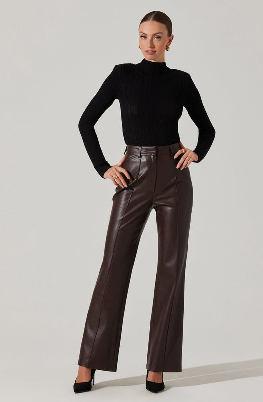 A woman is wearing a fitted, long-sleeved black ribbed turtleneck sweater, paired with high-waisted, straight-legged brown leather pants. She has on black high-heeled shoes and accessorizes with gold earrings and rings.