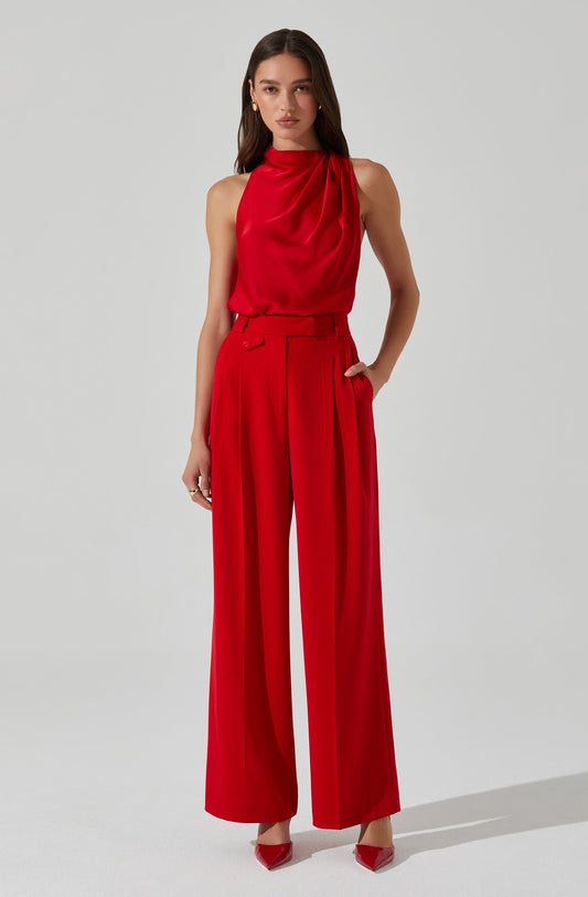 A woman is wearing a sleeveless, high-neck red blouse with draped fabric at the neckline, paired with high-waisted, wide-leg red trousers with pleats and a matching belt. She completes the outfit with red pointed-toe shoes.