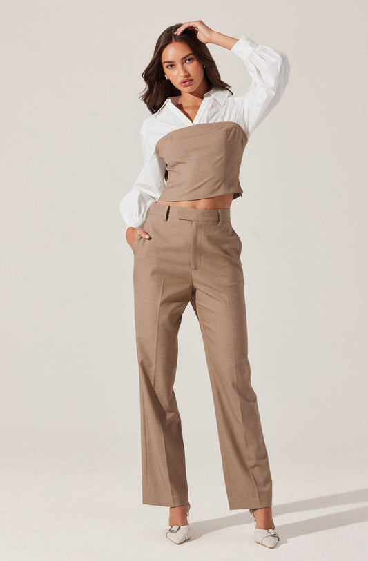 A woman is wearing a white long-sleeve blouse with a collar and puffed sleeves. Over the blouse, she has on a tan-colored strapless crop top. She is also wearing matching tan-colored high-waisted, straight-leg trousers with belt loops and front pockets. She has paired her outfit with white pointed-toe shoes.