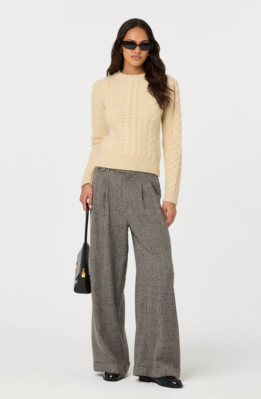 A woman is wearing a cream-colored, cable-knit sweater with long sleeves and a round neckline. She has on wide-leg, gray plaid trousers with a high waist and front pleats. She accessorizes with dark sunglasses, and is holding a black handbag with a gold clasp in her right hand.