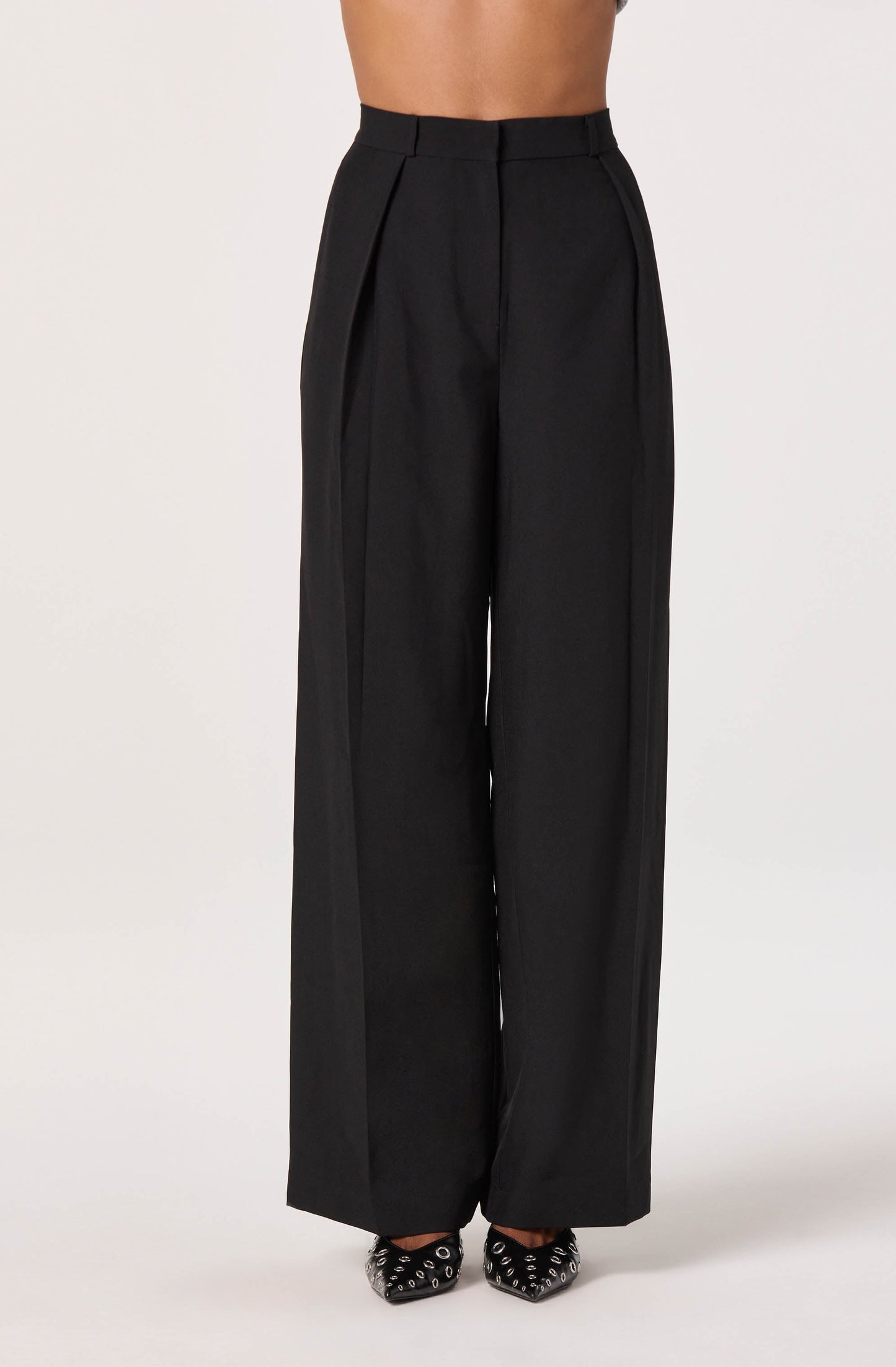 High-waisted black wide-leg trousers with pleats at the waist and subtle vertical seam detailing. The fabric appears to be structured and smooth. The person wearing the trousers is also wearing black pointed-toe shoes with silver eyelet embellishments.