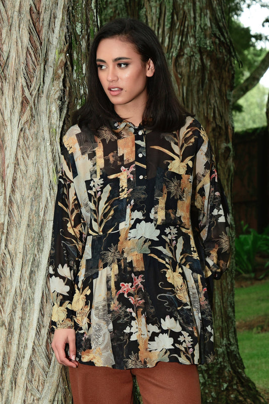 Model wears a long sleeve button-through shirt with a painterly floral print in earthy tones of gold, cream, rust and black. The shirt features a relaxed silhouette with a slight gathered waist and a classic collar. Styled with warm brown trousers, the look is set against a natural tree-lined backdrop for an effortless, organic feel.