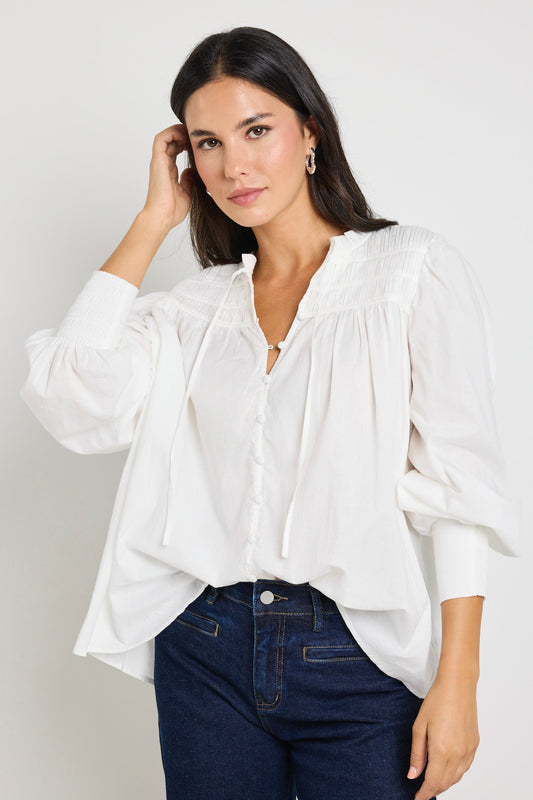 A woman wearing an ivory blouse with pintuck detailing, button-front closure, and tie neckline, styled with dark indigo mid-rise jeans.
