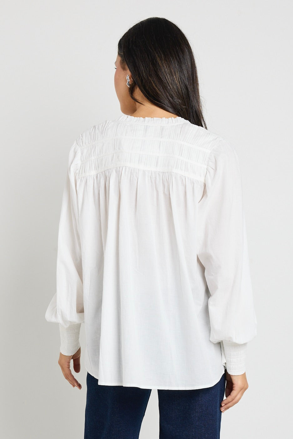 A woman wearing an ivory blouse with pintuck detailing, button-front closure, and tie neckline, styled with dark indigo mid-rise jeans.