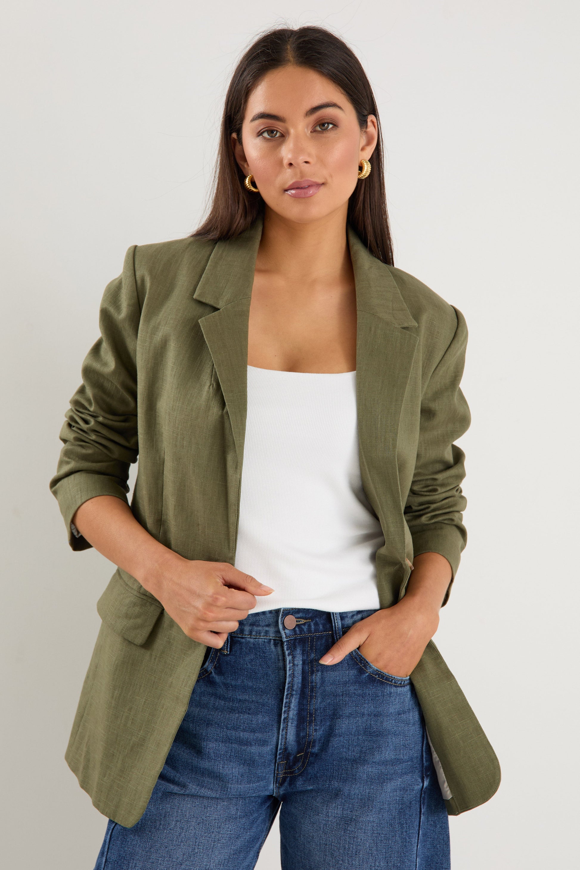 Woman wearing an olive green blazer over a white top and blue jeans on a plain background